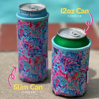 Two vibrant, floral-patterned Emerson West Resort Blooms Neoprene Can Coolers are shown by a poolside, perfect for poolside relaxation. The left cooler fits a 12 oz slim can while the right cooler holds a standard 12 oz can. Both coolers boast colorful, intricate designs with visible text indicating their respective can sizes.