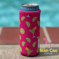 A slim neoprene can cooler adorned with a pink pattern featuring lime slices, palm leaves, and margarita glasses with umbrellas keeps a Blue Moon Brewing Co. 12 oz can cold. The can is placed on a stone ledge in front of a swimming pool. A "Margarita Neoprene Slim Can Cooler by Emerson West" label is present in the bottom right.