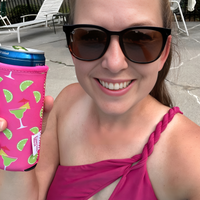 A woman wearing sunglasses and a twisted strap pink swimsuit smiles at the camera while holding a 12 oz drink in an Emerson West Margarita Neoprene Slim Can Cooler adorned with cocktail, lime slice, and palm leaf designs. She appears to be at a poolside with lounge chairs and a fence in the background.