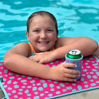 A young girl in a pool, leaning on the edge with her arms crossed on a pink and blue mat. She is smiling and holding a canned drink in one of those vibrant Flamingo Floats Neoprene Can Coolers by Emerson West. The background showcases the clear blue water sparkling under the sun.