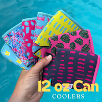 A hand fan is holding several colorful Emerson West Palm Leaves Neoprene Can Coolers with various patterns, including tropical leaves, abstract designs, and geometric shapes. The background shows a blurred swimming pool, and the text "12 oz Can Coolers" appears at the bottom.