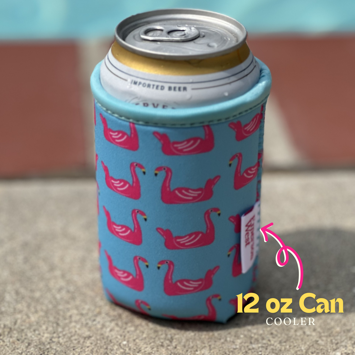 An Emerson West Flamingo Floats Neoprene Can Cooler with blue fabric featuring pink flamingos holds a silver beer can. A pink arrow and text state "12 oz Can Cooler." The background is an outdoor setting with concrete and an out-of-focus pool edge.