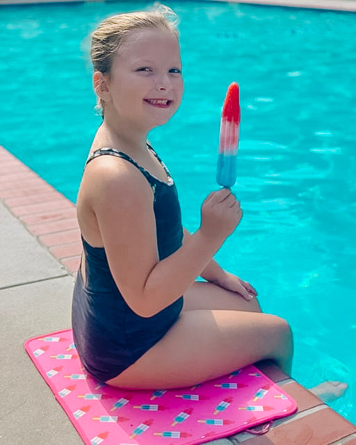 A young girl in a swimsuit enjoying a colorful Bomb Pops popsicle by the poolside on a sunny day, seated on an Emerson West neoprene pool mat.