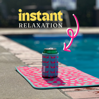 A 12 oz can of drink with a Palm Leaves Neoprene Can Coolers by Emerson West featuring pink flamingos stands on a mat with palm leaves and pink polka dots by the pool. Pink accents and an arrow point towards the can from text at the top that reads "Instant Relaxation." The background is slightly blurred.