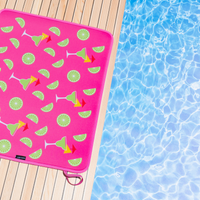 A vibrant pink, quick-dry Margaritas Poolside Seating Mat decorated with lime and cocktail graphics resting on a wooden deck next to a shimmering blue pool. From Emerson West.