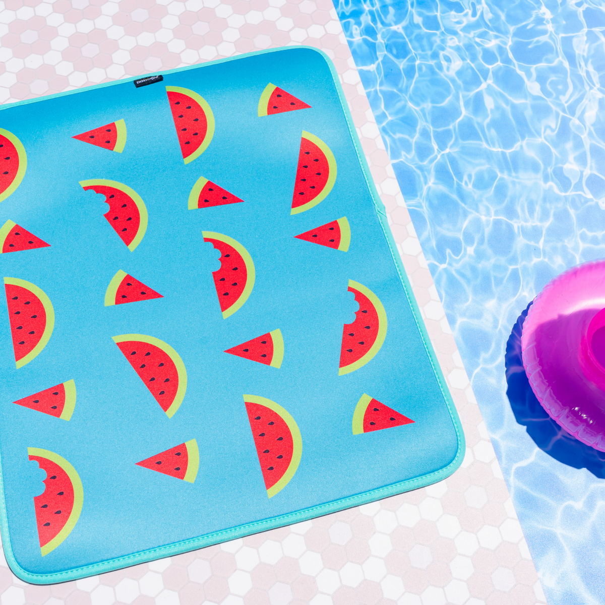 A vibrant blue, fade-resistant Emerson West Watermelon Poolside Seating Mat adorned with watermelon slices near the edge of a swimming pool, complementing the bright pink swim ring bobbing on the sparkling water under a sunny sky.