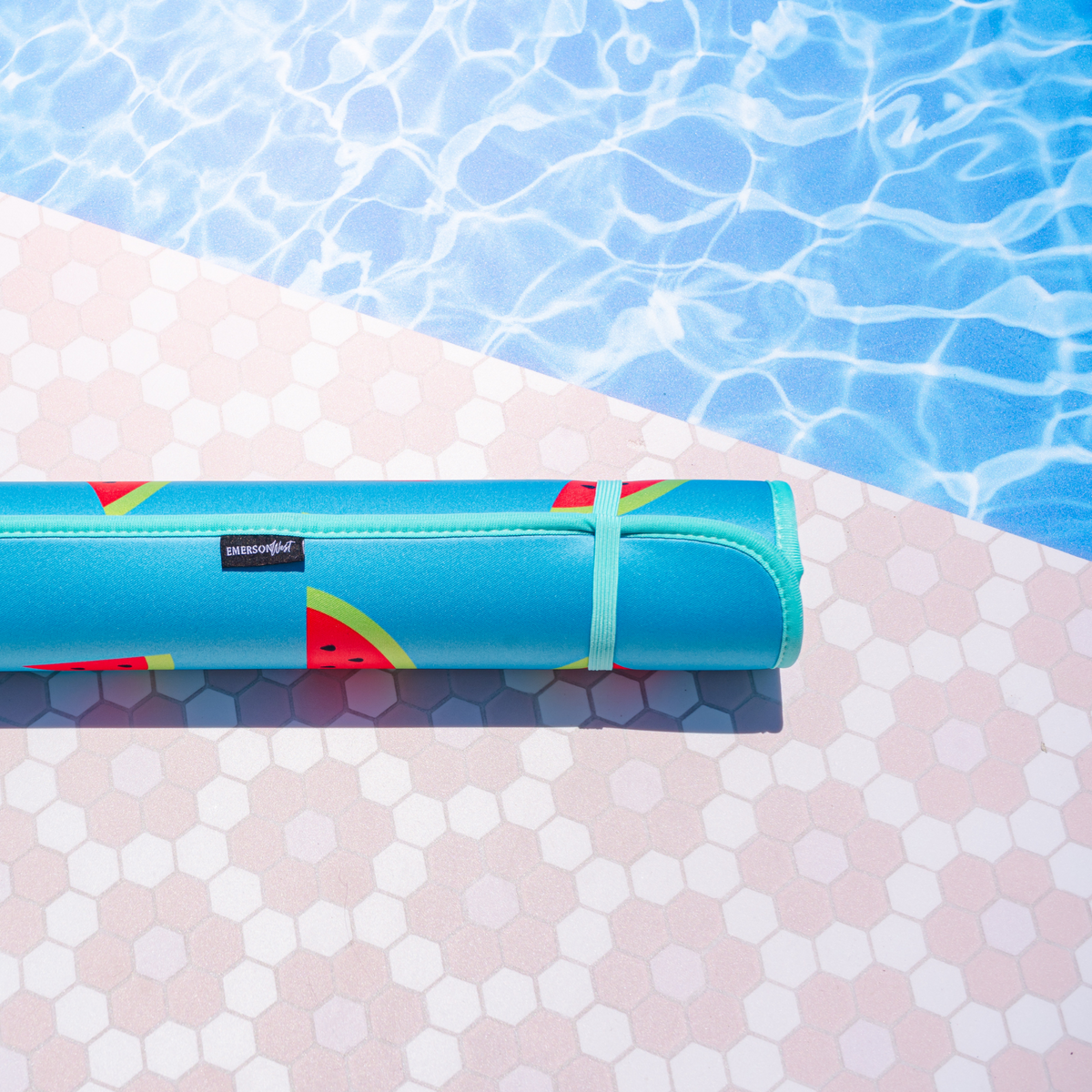 A bright blue, quick-dry Emerson West Watermelon Poolside Seating Mat rests on the edge of a pool, with sun-dappled water shimmering in the background and pink and white tiles in the foreground.