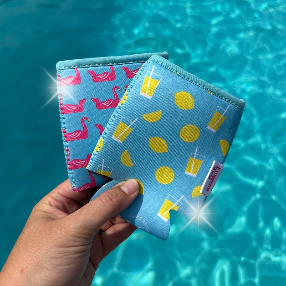 A hand holds two colorful 12 oz can coolers against a backdrop of a shimmering blue pool. The sleeve on the left features Emerson West's Flamingo Floats Neoprene Can Cooler with pink flamingos on a light blue background, while the one on the right has yellow lemons and glasses of lemonade on a matching neoprene background.