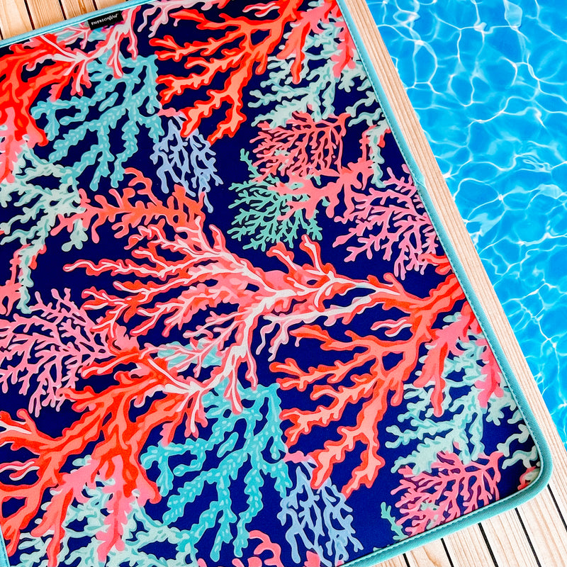 A colorful, fade-resistant Coral Poolside Seating Mat by Emerson West adorned with a vibrant coral reef pattern in shades of red, pink, blue, and green lays on a wooden deck next to a clear, bright blue swimming pool.