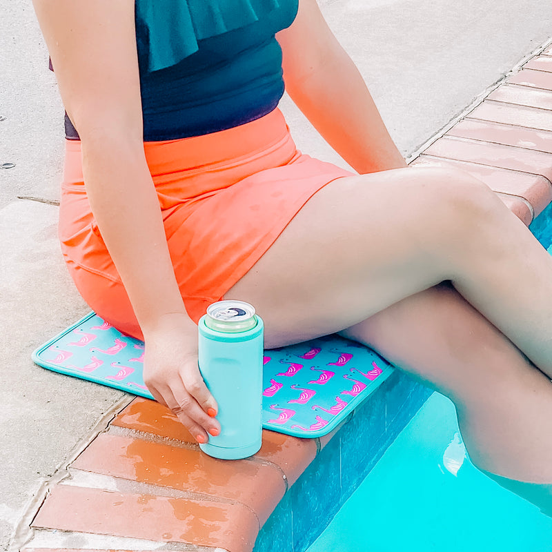 Woman sitting poolside on a sunny day, dressed in a vibrant orange and black swimsuit, enjoying a cold beverage poolside while sitting on an Emerson West poolside mat. Enjoying some leisure time by the pool while also protecting her swimsuit and skin.