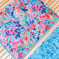 A vibrant, multicolored floral and leaf-patterned, quick-dry mat lies on a wooden deck adjacent to a blue swimming pool. The Emerson West Resort Blooms Poolside Seating Mat features swirling designs in shades of pink, blue, green, and orange, adding a lively, tropical feel to the setting.