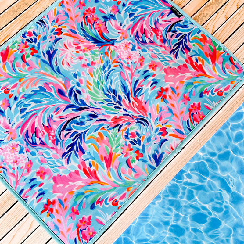 A vibrant, multicolored floral and leaf-patterned, quick-dry mat lies on a wooden deck adjacent to a blue swimming pool. The Emerson West Resort Blooms Poolside Seating Mat features swirling designs in shades of pink, blue, green, and orange, adding a lively, tropical feel to the setting.