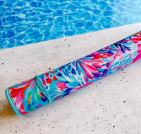 An Emerson West Resort Blooms Poolside Seating Mat with a colorful floral design, featuring shades of pink, blue, and purple, is rolled up and placed on a stone surface. In the background, clear blue water of an outdoor poolside is visible. This fade-resistant mat is perfect for quick-dry use after a swim.