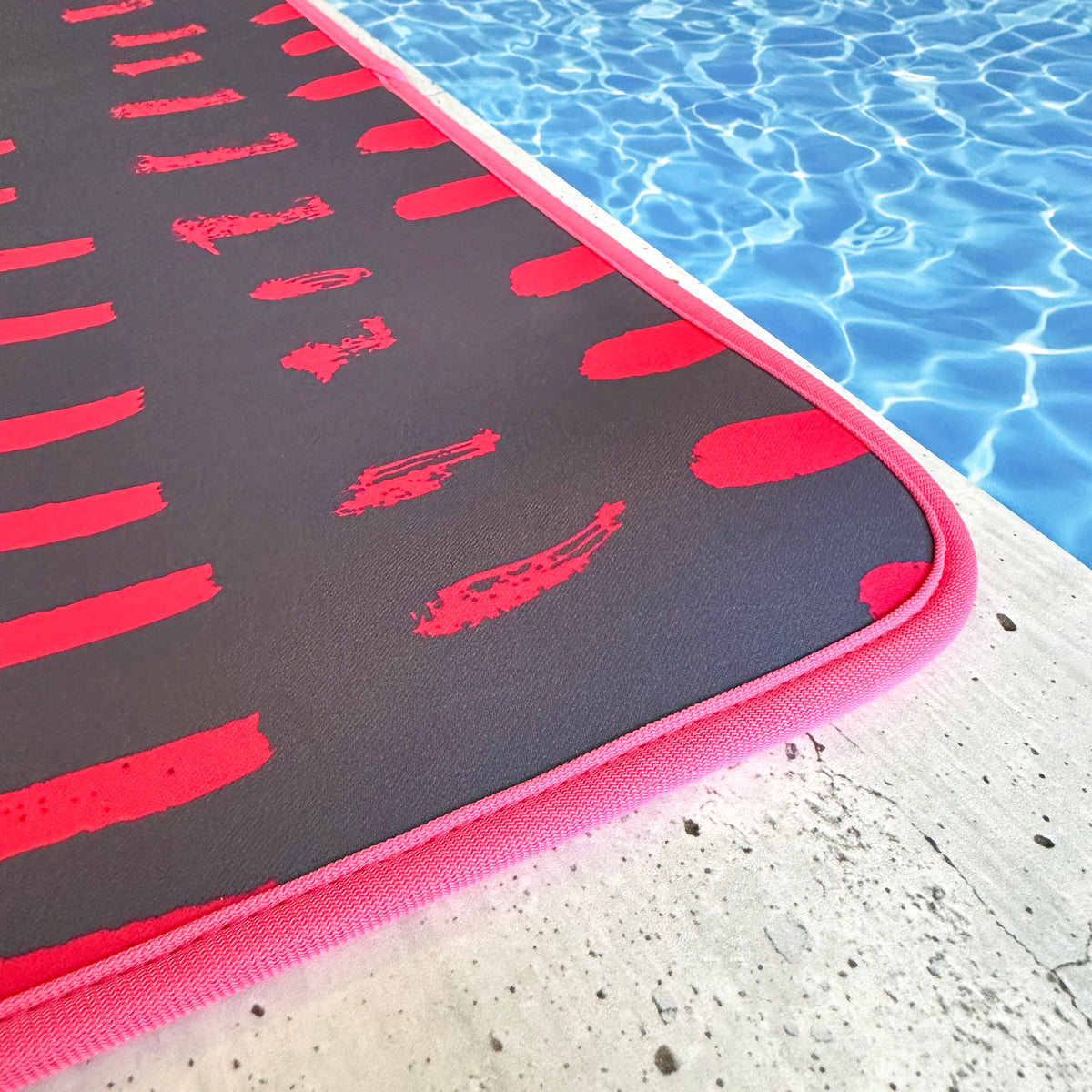 A Navy & Pink Poolside Seating Mat by Emerson West rests on the edge of a swimming pool. This poolside seating mat features a red border, while the clean and inviting water seems ideal for relaxation. Plus, it's quick-dry and fade-resistant, perfect for any outdoor setting.