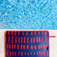 A colorful, fade-resistant Navy & Pink Poolside Seating Mat by Emerson West with a pattern of red and orange vertical dashes and a red trim is placed on a light-colored concrete surface. The quick-dry mat is near a clear blue swimming pool, with the pool water’s ripples creating a shimmering effect.