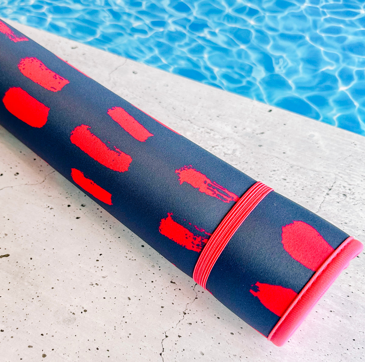 A rolled-up yoga mat with red and pink patterns lies on the edge of a concrete surface next to a blue swimming pool. This fade-resistant Emerson West Navy & Pink Poolside Seating Mat, secured with a pink elastic strap, doubles as quick-dry poolside seating.