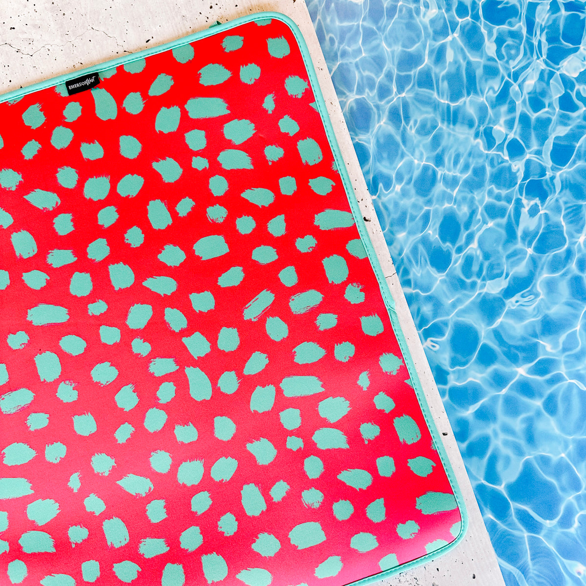 A vibrant pink and turquoise-patterned, fade-resistant Emerson West Pink Cheetah Poolside Seating Mat resting by the glistening blue waters of a swimming pool, inviting relaxation under the sun.