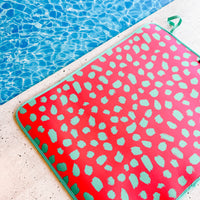 A vibrant Pink Cheetah Poolside Seating Mat with a fade-resistant teal cheetah pattern perched on the pool deck overlooking  the clear blue water of a swimming pool, hinting at leisure and summertime fun by Emerson West.