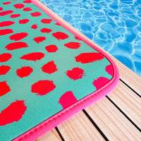 A vibrant Teal Cheetah Poolside Seating Mat from Emerson West with a hot pink cheetah pattern and a non-slip bottom rests on a wooden deck beside a serene, sunlit swimming pool.