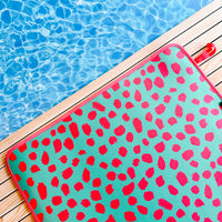 A vibrant teal and pink cheetah patterned quick-dry Teal Cheetah Poolside Seating Mat laid out on a wooden deck beside a sparkling swimming pool, inviting a refreshing summer day dip by Emerson West.