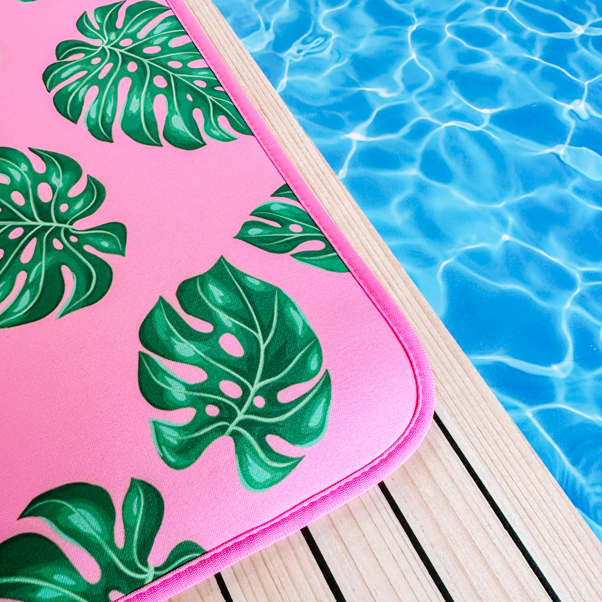 A vibrant pink pool mat with a tropical Palm Leaves Poolside Seating Mat design by Emerson West, resting on a wooden deck beside the rippling blue waters of a swimming pool, evoking a sense of summertime relaxation.