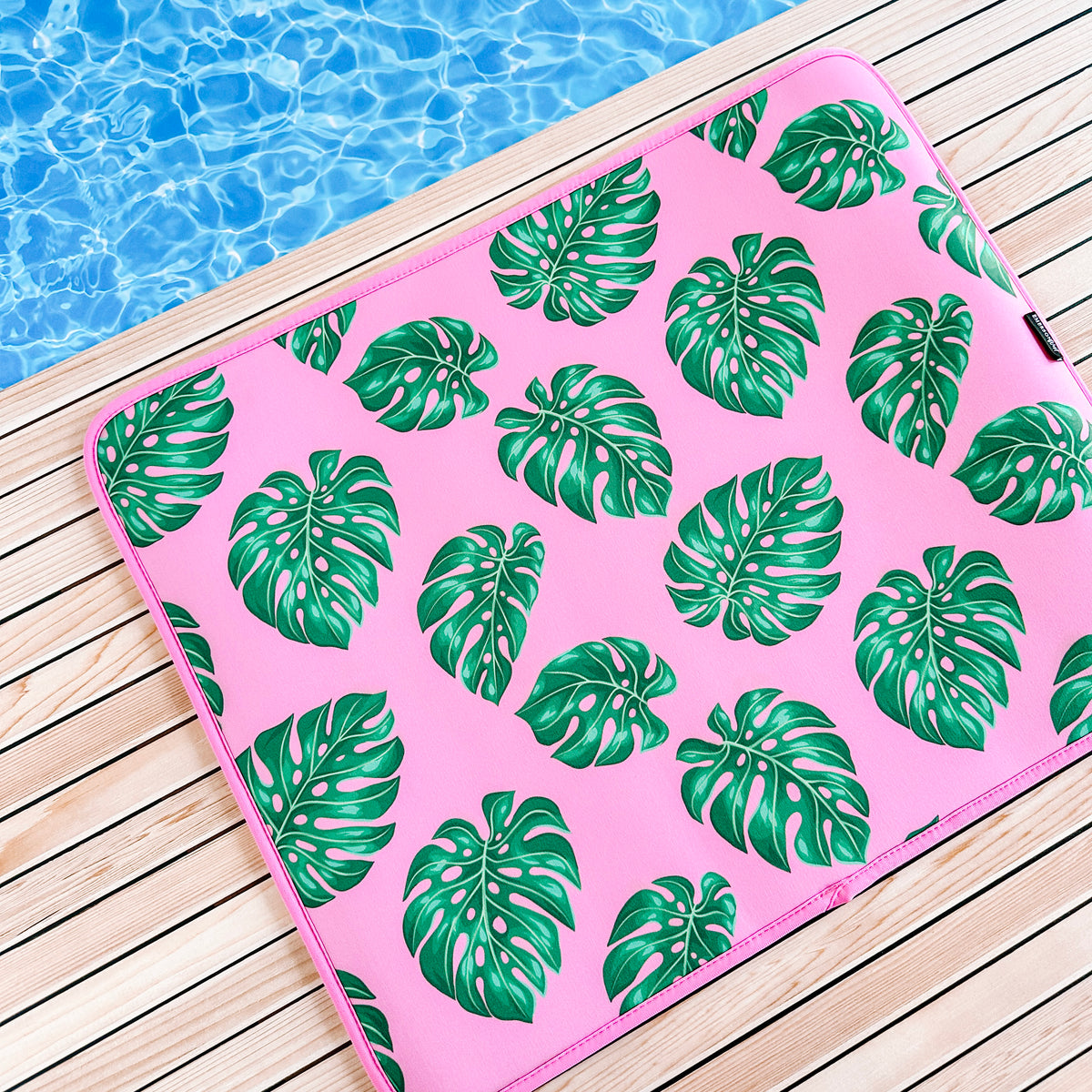 A vibrant pink poolside seat cushion. The Palm Leaves Poolside Seating Mat by Emerson West sitting on a wooden deck beside a pool with clear blue water, on a quick-dry, fade-resistant poolside mat.