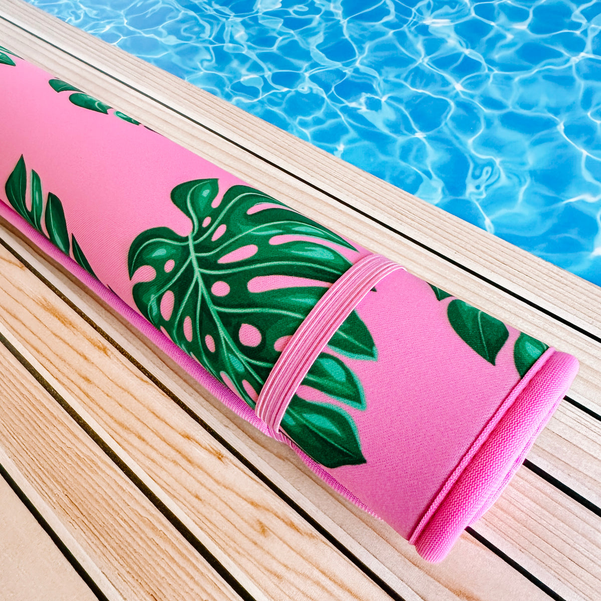 A fade-resistant pink Palm Leaves Poolside Seating Mat adorned with a tropical palm leaf design rests on a wooden deck beside a sparkling blue swimming pool. (Brand name: Emerson West)