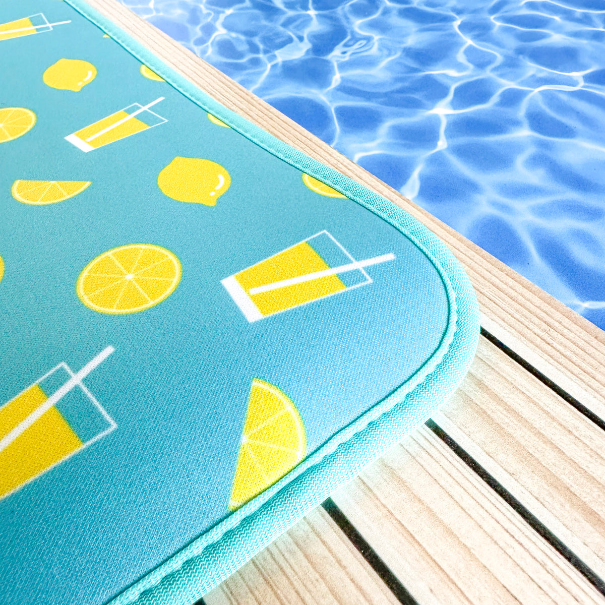 A vibrant aqua-colored neoprene Lemonade Poolside Seating Mat with lemon and drink illustrations sits beside the shimmering blue water of a swimming pool, evoking a sense of summertime relaxation by Emerson West.