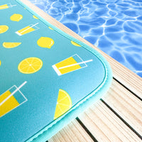 A vibrant aqua-colored neoprene Lemonade Poolside Seating Mat with lemon and drink illustrations sits beside the shimmering blue water of a swimming pool, evoking a sense of summertime relaxation by Emerson West.