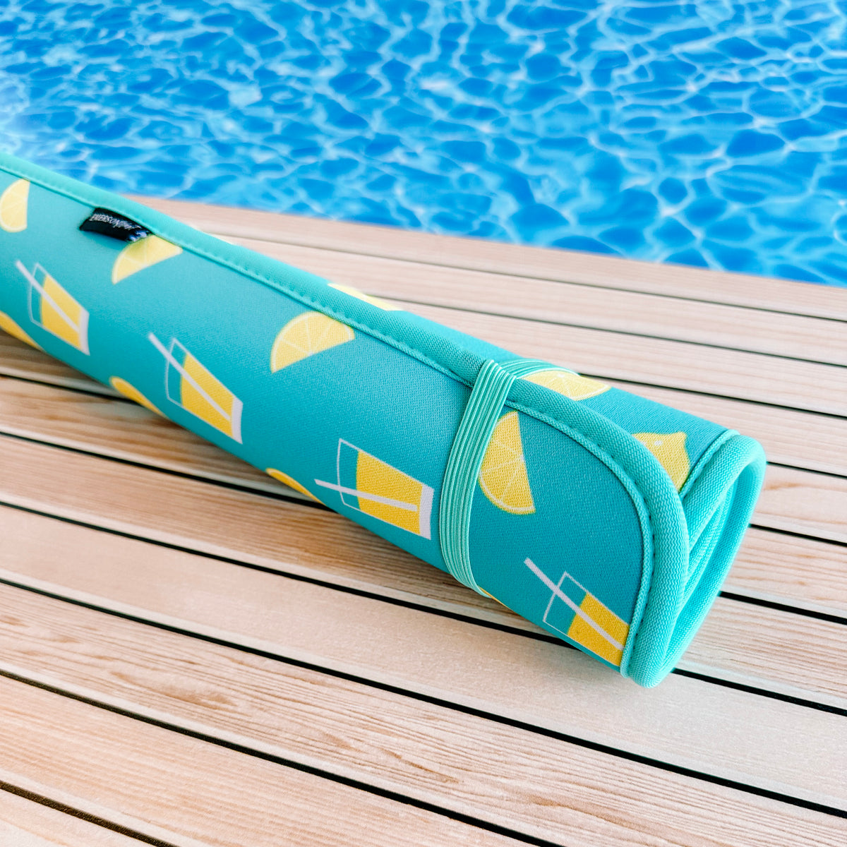 A Lemonade Poolside Seating Mat by Emerson West with a lemon and beverage glass design rolled up and resting on a wooden deck by a sparkling blue pool.