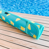 A Lemonade Poolside Seating Mat by Emerson West with a lemon and beverage glass design rolled up and resting on a wooden deck by a sparkling blue pool.