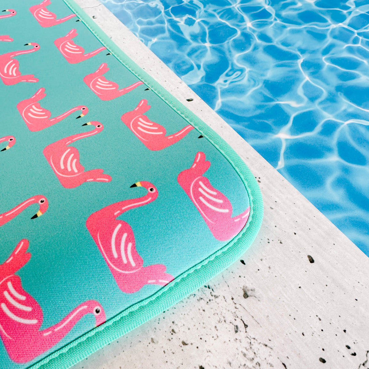 A vibrant turquoise Emerson West poolside mat adorned with vivid pink Flamingo Floats patterns resting on a concrete edge beside the tranquil, shimmering waters of a swimming pool.