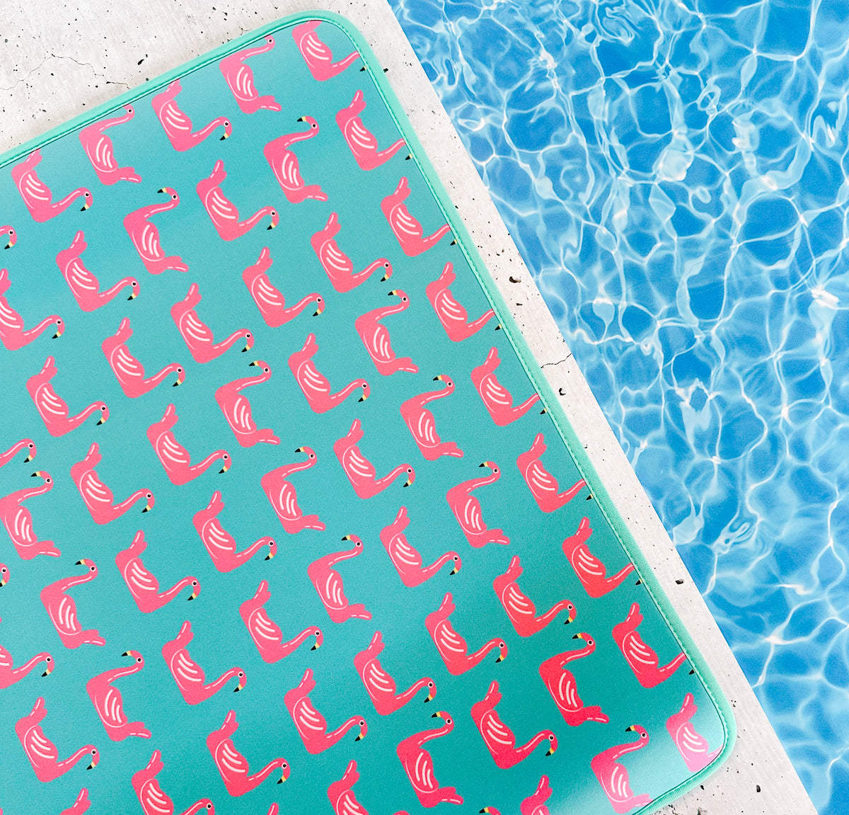 A vibrant aqua-colored Emerson West Flamingo Floats Poolside Seating Mat adorned with a pattern of pink flamingos floats serenely on the shimmering blue water of a swimming pool, suggesting a fun and relaxing summertime.