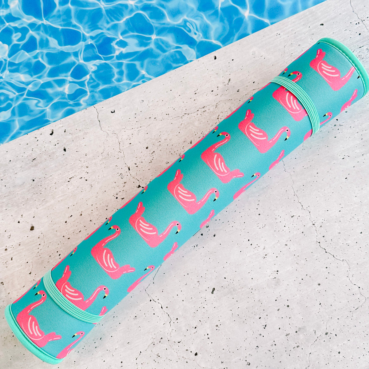 A Flamingo Floats Poolside Seating Mat by Emerson West with a pattern of pink flamingos, beside a swimming pool with clear blue water.