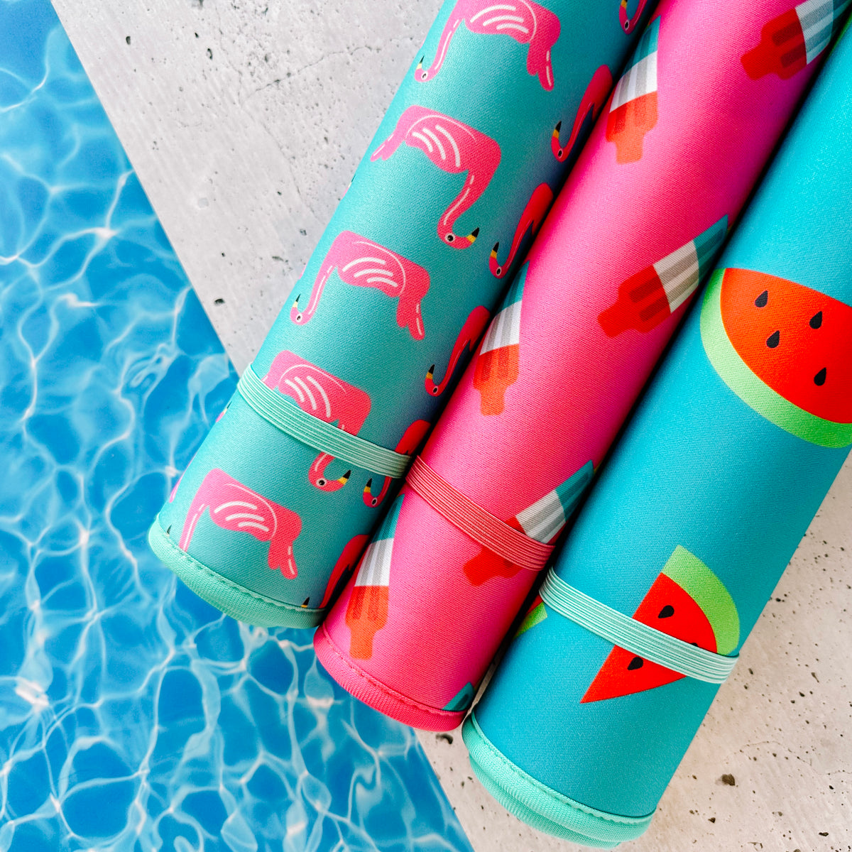 Colorful Pool Party Collection pool accessories featuring flamingo floats, popsicles, and watermelon prints resting by the edge of a swimming pool, poolside must-haves. Brand Name: Emerson West