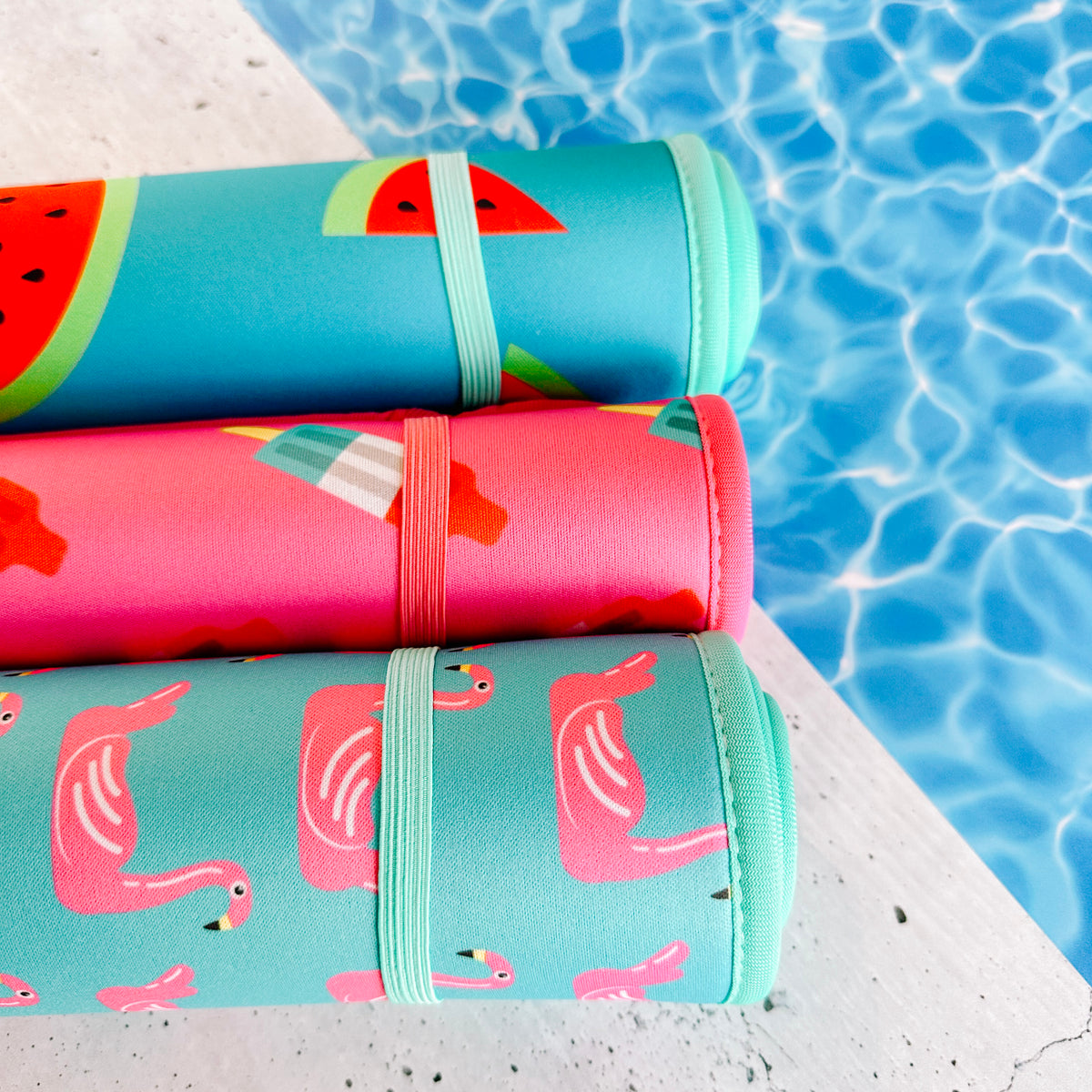 Colorful neoprene Pool Party Collection mats with whimsical watermelon and flamingo designs from Emerson West, ready for a fun day by the sparkling blue water of a swimming pool.