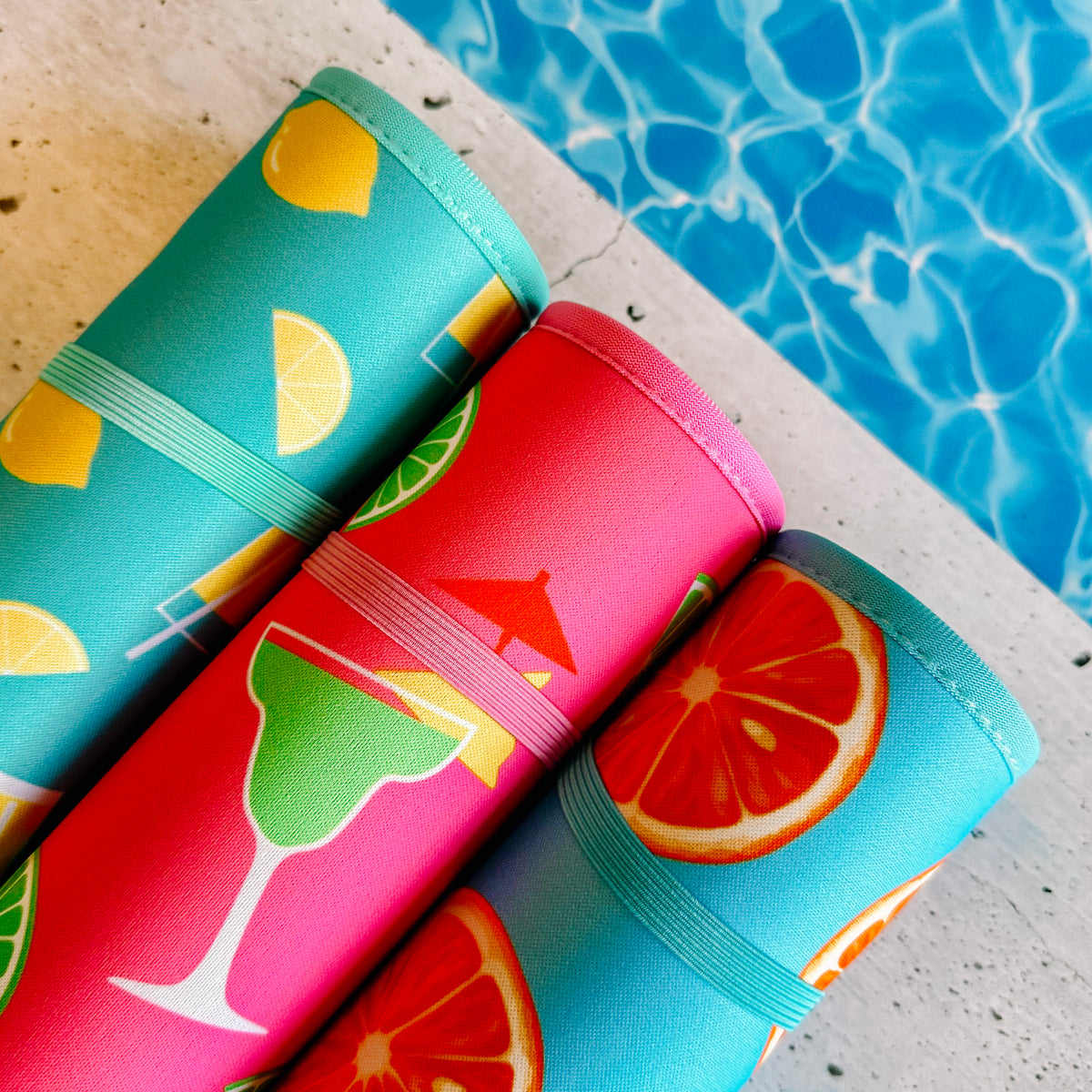 Fade-Resistant, colorful patterned poolside seating mats from the Citrus Delight Collection by Emerson West, with citrus fruit designs, arranged at the edge of a swimming pool with clear blue water.