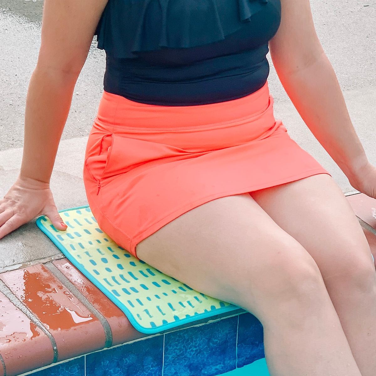 A person sitting by the poolside on an Emerson West neon yellow poolside seating mat wearing a black top and a bright orange swim skirt, with their legs partially in the water.