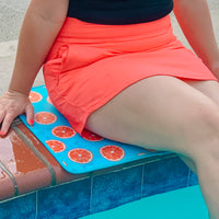 A person sitting poolside with their legs partially dipped in the water, resting on a colorful, quick-dry neoprene poolside mat with an oranges design by Emerson West.