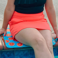 A person in a black top and an orange skirt sits at the edge of a swimming pool, with their legs partially resting on an Emerson West Oranges Poolside Seating Mat with citrus fruit designs.