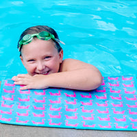 A joyful child with swim goggles rests on an Emerson West Flamingo Floats Poolside Seating Mat adorned with pink flamingo patterns in a bright blue swimming pool.