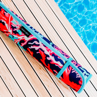 A rolled-up Coral Poolside Seating Mat with a vibrant red, blue, and white coral pattern lies on a wooden pool deck. The blue water of the pool contrasts with the colorful, fade-resistant mat by Emerson West, which has a teal strap securing it.