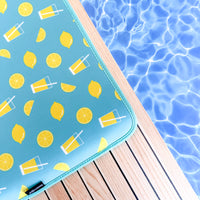 A vibrant turquoise pool float from the Citrus Delight Collection by Emerson West with a lemon and beverage print resting on a wooden deck beside a shimmering blue pool, featuring fade-resistant material.