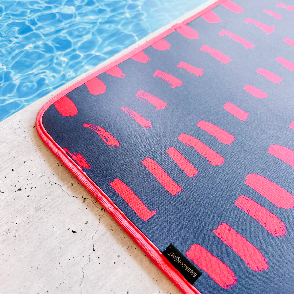 A Navy & Pink Poolside Seating Mat with a pink and blue abstract design lies next to a pool with clear blue water. This quick-dry mat, perfect for poolside seating, has a tag labeled "Emerson West." The pool area is bordered by light-colored concrete.