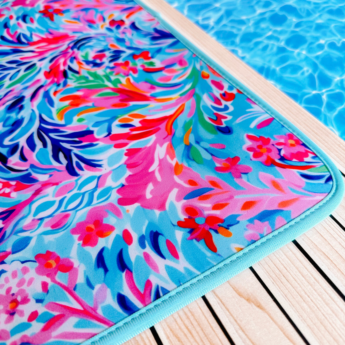 A close-up shot of a colorful, floral Resort Blooms Poolside Seating Mat by Emerson West with pink, blue, and red patterns lying on a wooden deck near a swimming pool. The fade-resistant mat's vibrant design contrasts with the clear, blue water of the pool.