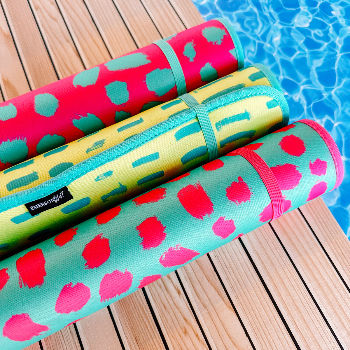 Colorful poolside mats from the Oh So Bright Collection by Emerson West rolled up on a wooden deck beside a glistening pool.