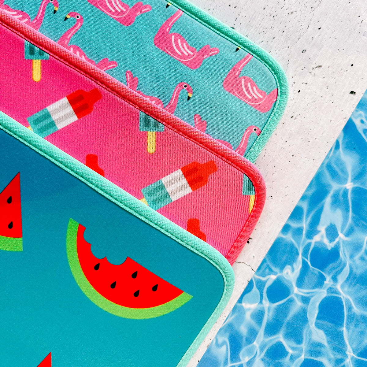 A trio of bright, tropical-themed Pool Party Collection poolside seating mats with flamingo, popsicle, and watermelon designs resting near a poolside, evoking a summery vibe by Emerson West.