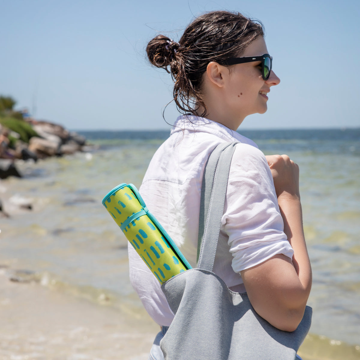 A woman enjoys a sunny day at the beach carrying a Neon Yellow Poolside Seating Mat in her bag, ready for a relaxing day by the sea. Brand name: Emerson West